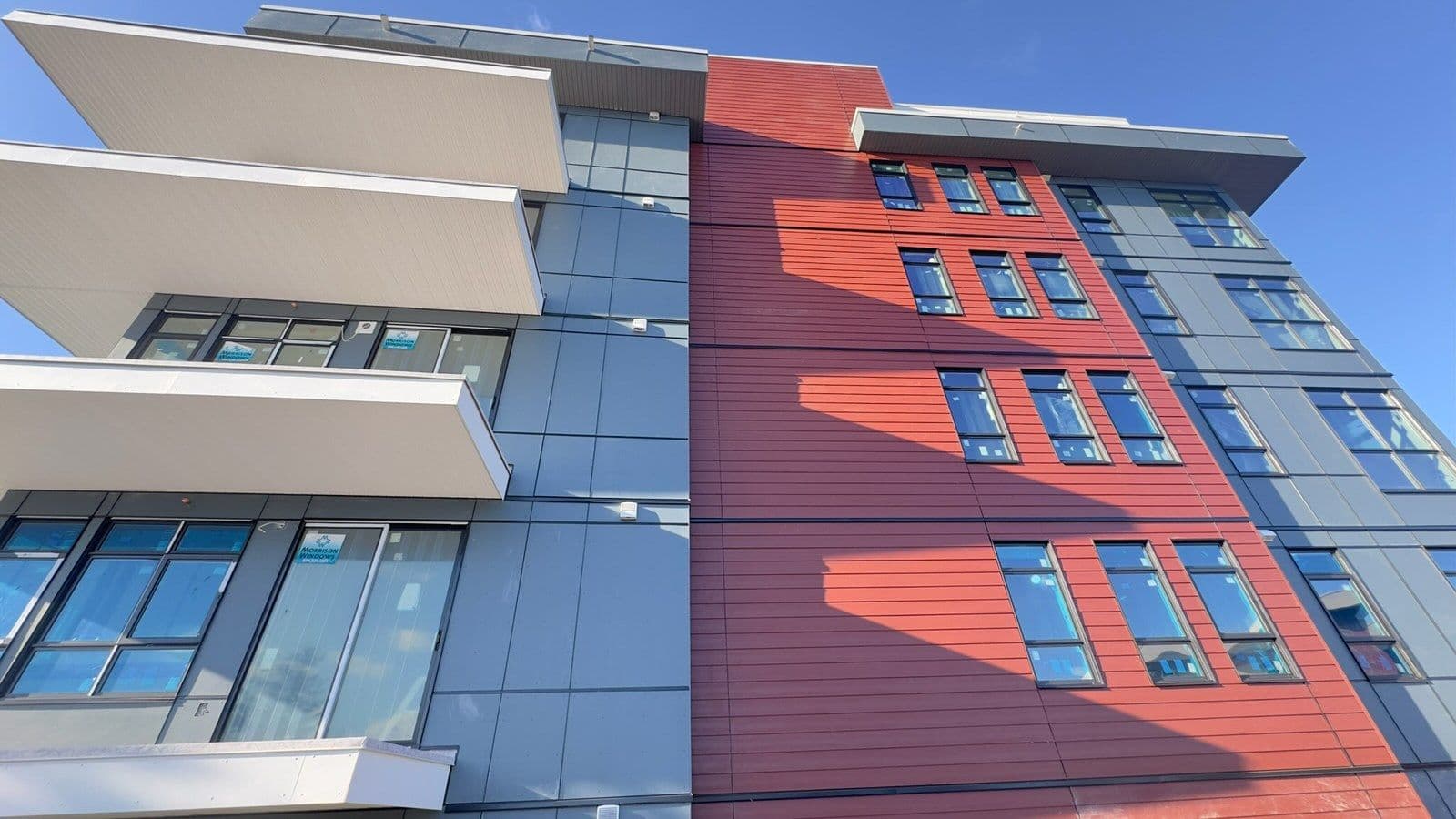Modern multi-storey building with red horizontal siding panels and blue-gray cladding, featuring rows of black-framed windows and protruding white balconies under a clear blue sky.