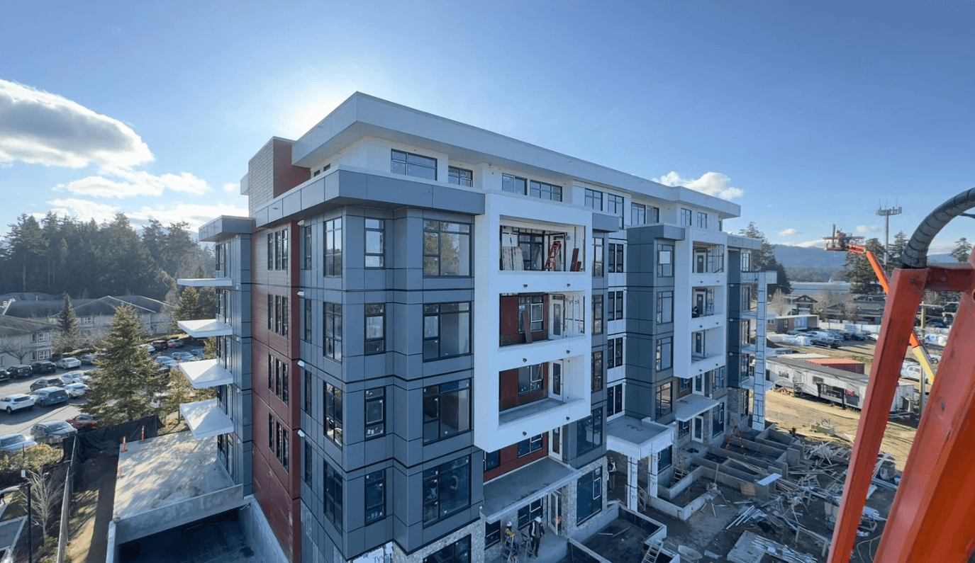 Modern mid-rise apartment building under construction with gray and white siding, large windows, and balconies, photographed on a clear day with trees and houses in the background.