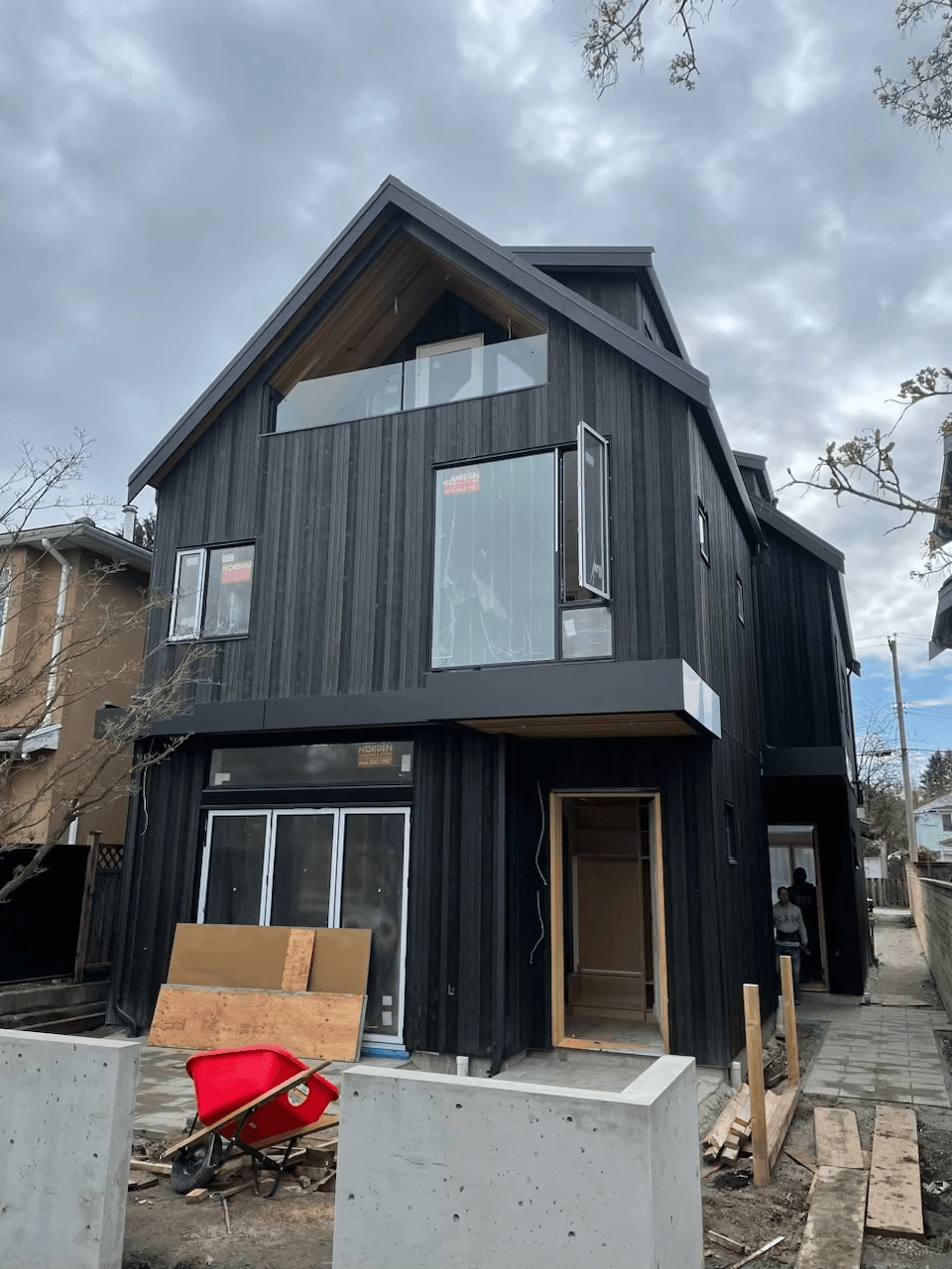 Contemporary black-sided house under construction with large windows and a red wheelbarrow in front